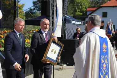 Dwaj mężczyźni w garniturach wręczają kapłanowi w białej szacie z niebieskim ornatem oprawiony dyplom z medalami podczas uroczystości plenerowej na scenie.